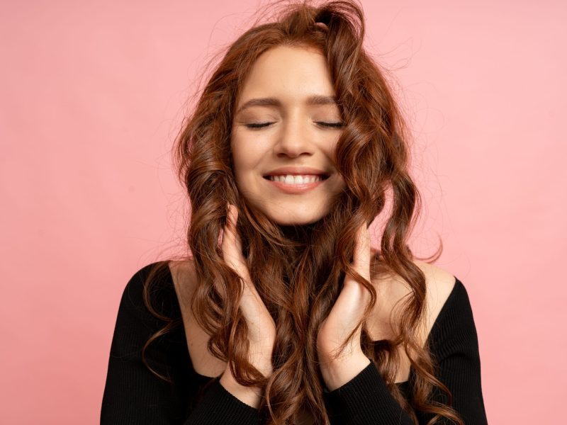 Beautiful red  head woman  with close eyes posing over pink background. Wavy hairs. Perfect smile.  Isolate in studio.